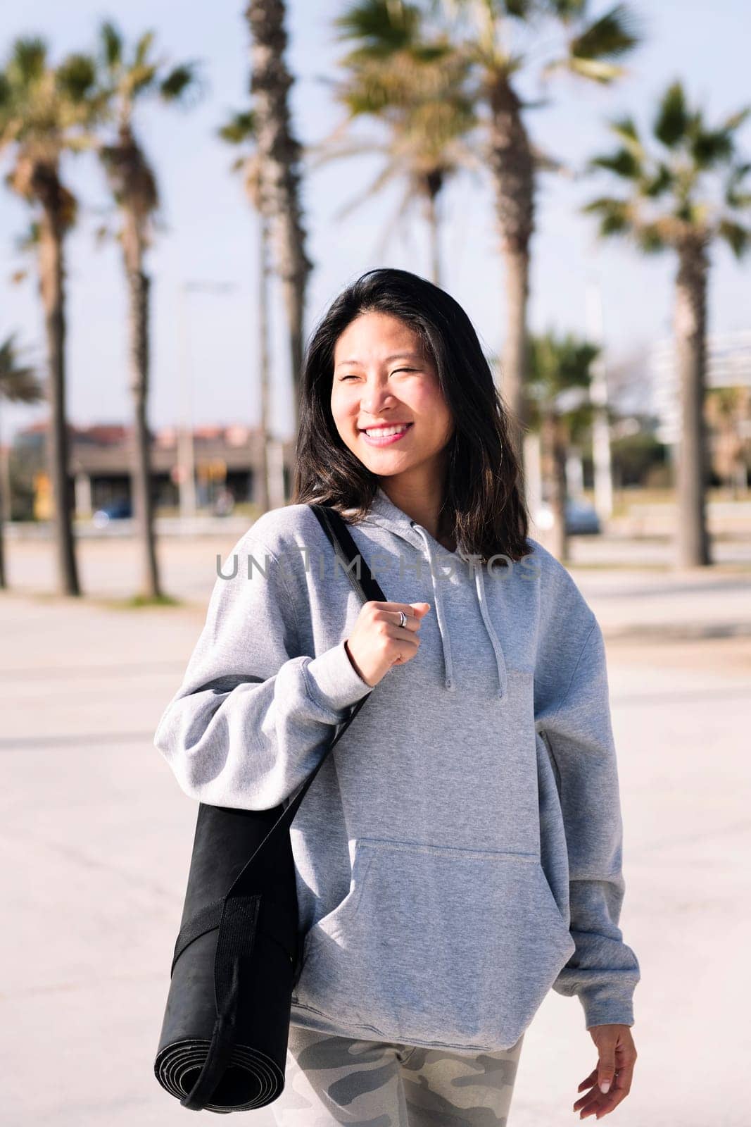 smiling young asian woman dressed in casual clothes walking with yoga mat hanging on her arm, sport and healthy lifestyle concept