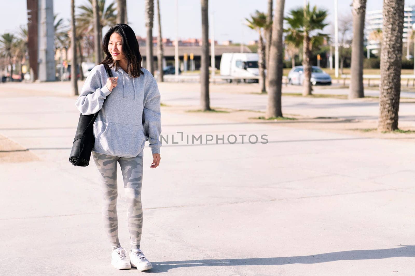 smiling young asian woman dressed in casual clothes walking with yoga mat hanging on her arm, sport and healthy lifestyle concept, copy space for text