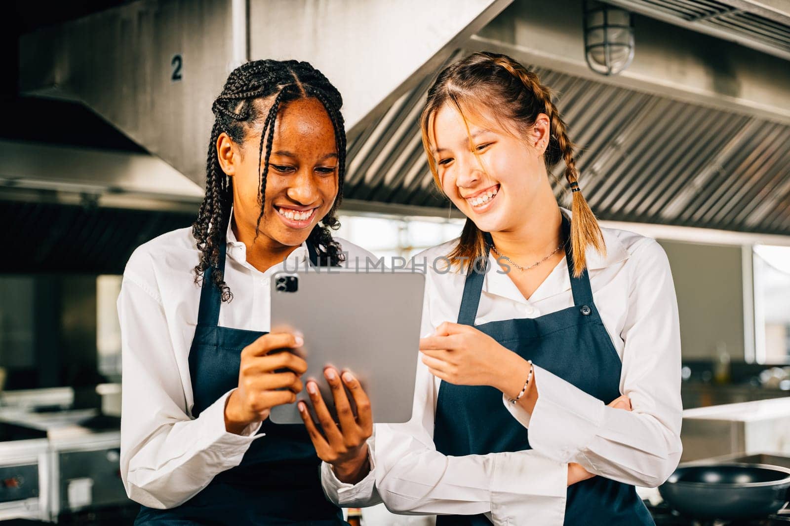 Two famous chefs discussing their video blog in a professional stainless steel kitchen using a tablet. Professionals smiling reading searching communicating with modern technology.