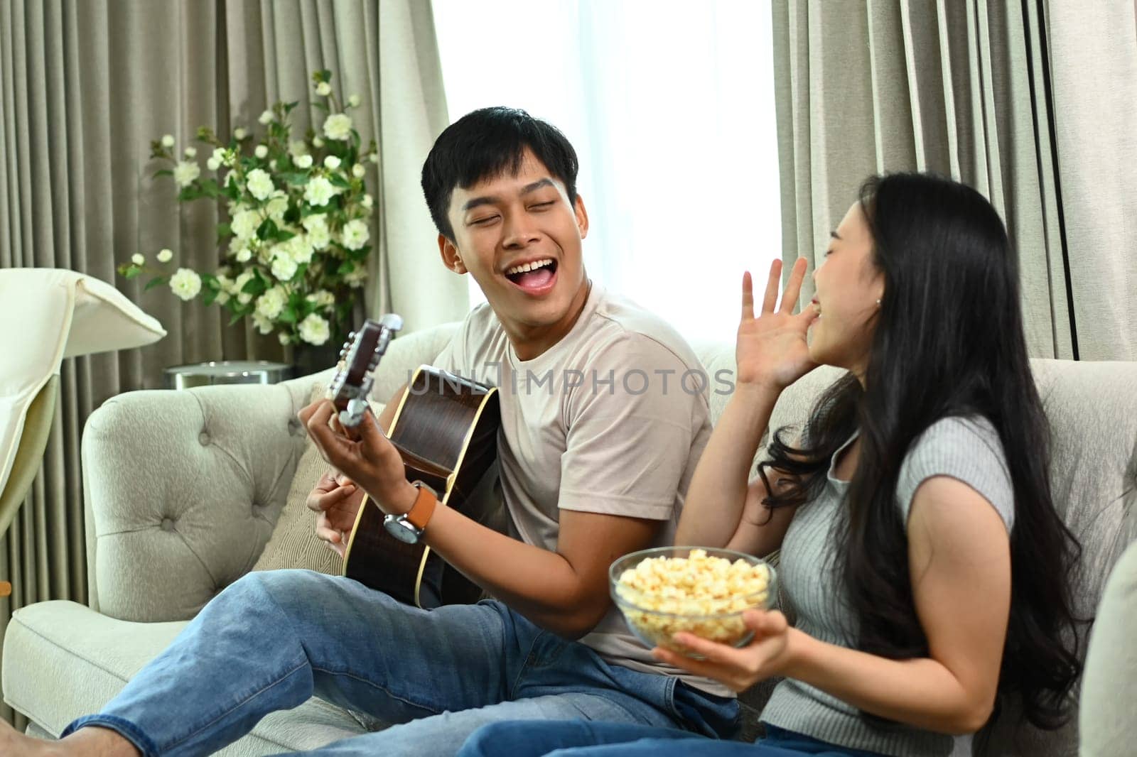 Shot of young man playing guitar for his girlfriend sitting on couch at home by prathanchorruangsak