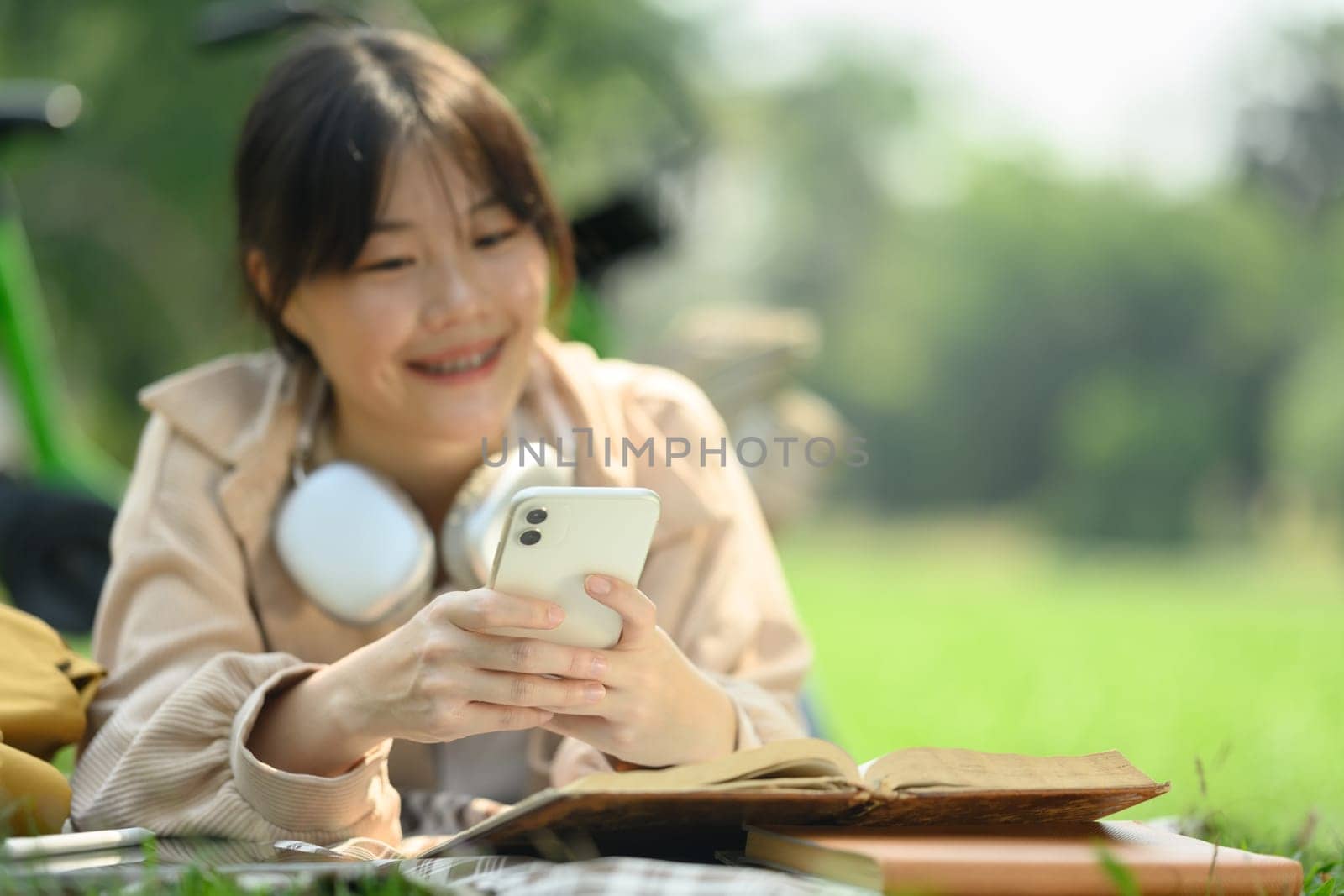 Delighted Asian young woman lying on blanket with books and using mobile phone.