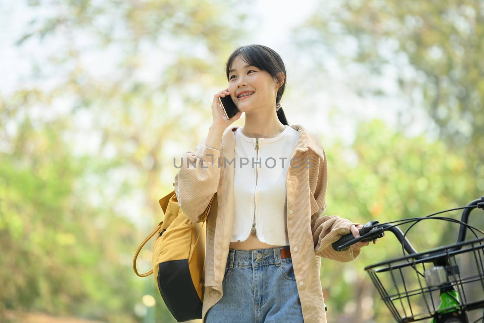 Positive young woman in casual clothes talking on mobile phone and standing near bicycle by prathanchorruangsak