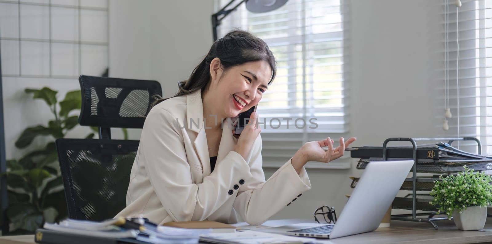 Young Asian business woman sits on the phone in an online business meeting using a laptop in a modern home office decorated with shady green plants..