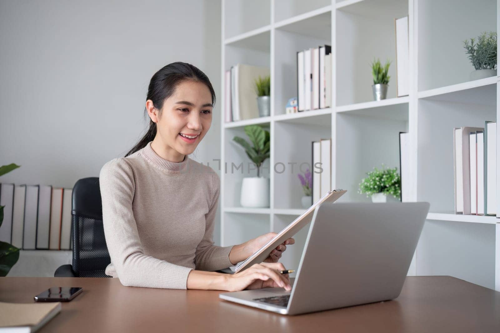 Asian businesswoman or accountant happily sitting and working with laptop on finance and business administration. On the desk in the office decorated with green plants..