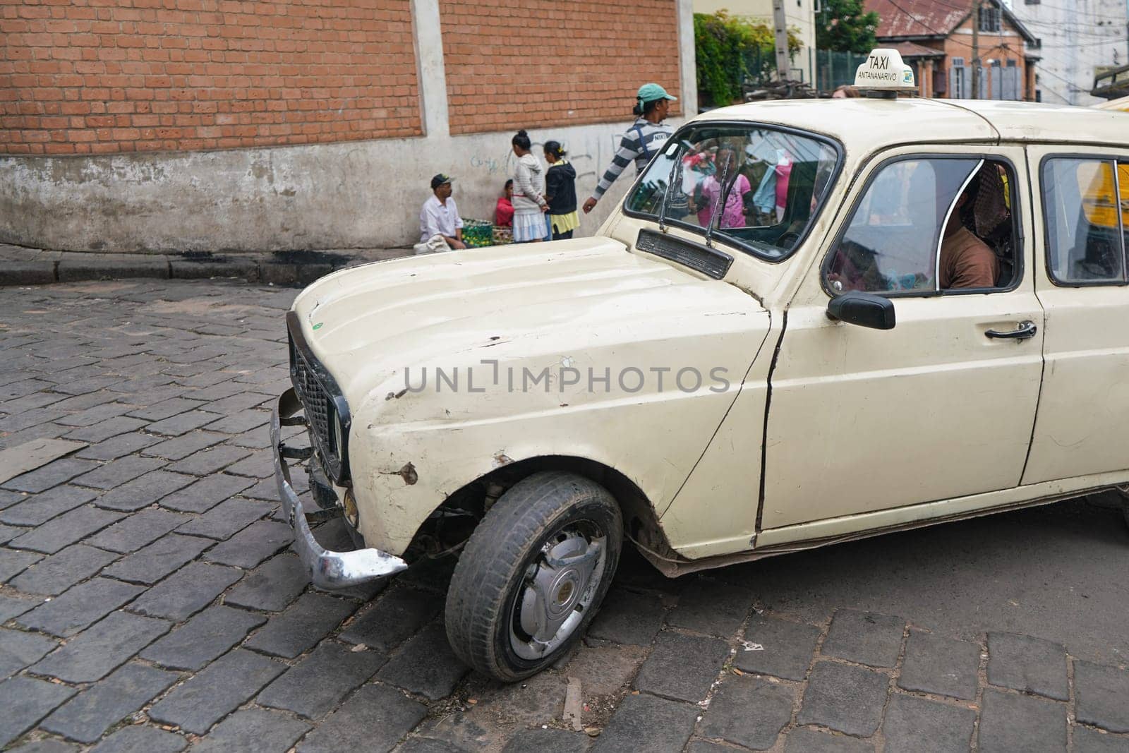 Antananarivo, Madagascar - April 24, 2019: White taxi car driving slowly over tile stone road, group of unknown Malagasy people in background. Madagascar is poor country, most vehicles are worn out
