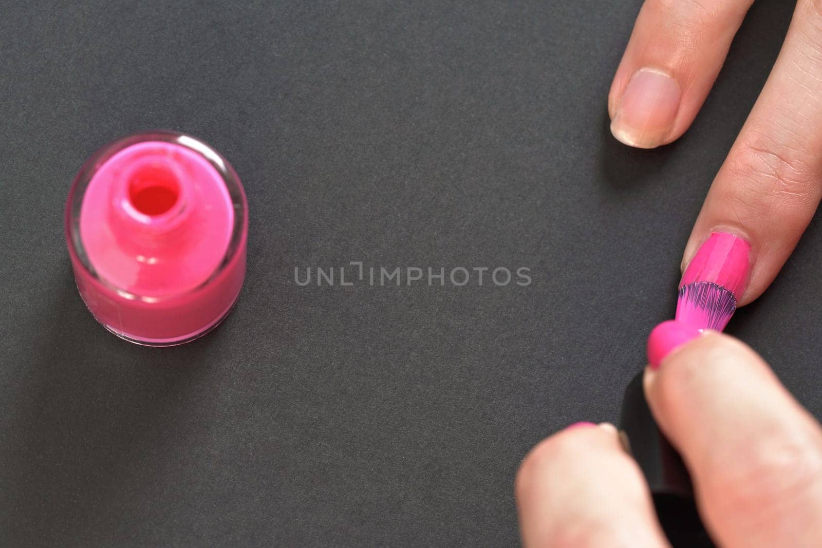 Applying pink nail polish - closeup photo of woman hands, bottle with varnish, little brush and fingernails on gray / black background by Ivanko