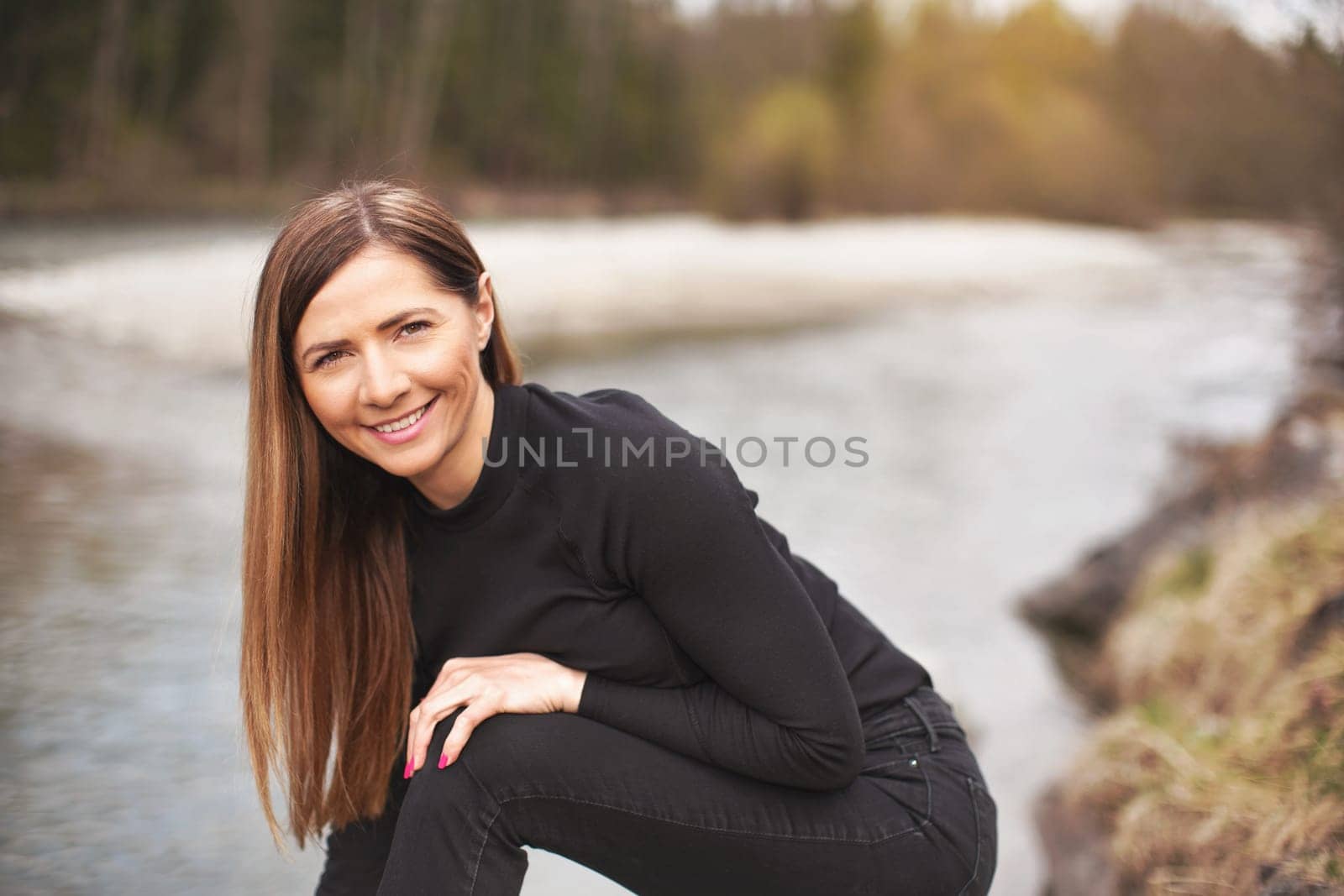 Portrait of young woman wearing black jeans and top, leaning, her long hair falling down, smiling. Blurred river in background