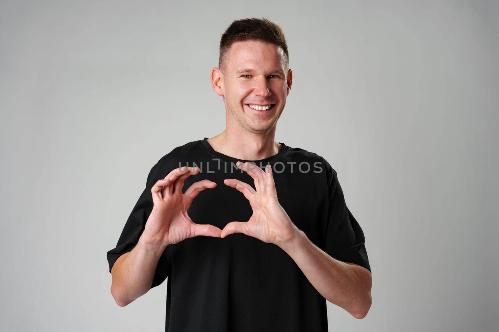 Smiling young man gesturing heart sign with ahnds against gray background close up