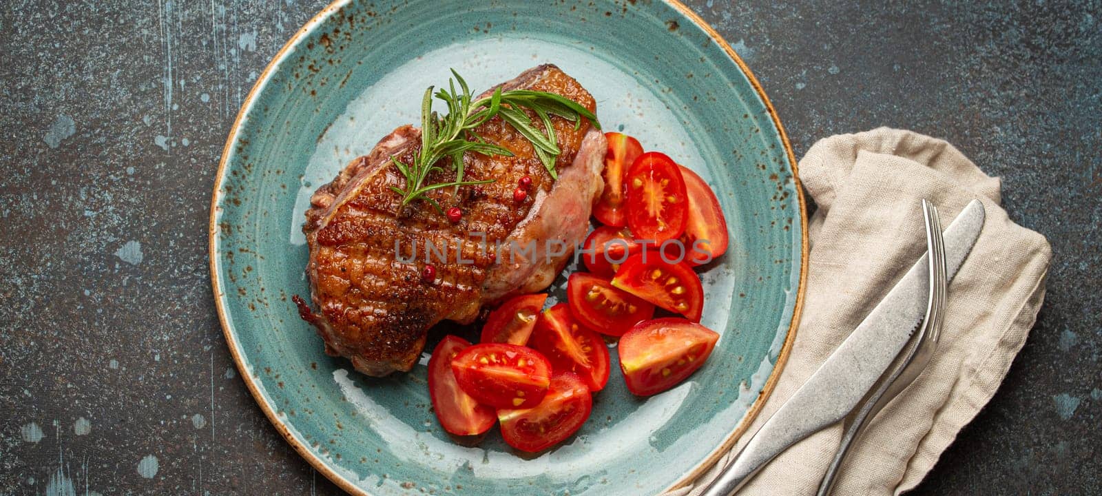 Delicious roasted duck breast fillet with golden crispy skin, with pepper and rosemary, top view on ceramic blue plate served with cherry tomatoes salad, rustic concrete rustic background by its_al_dente