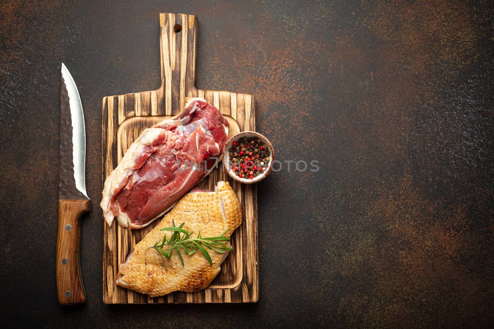 Two raw uncooked duck breast fillets with skin, seasoned with salt, pepper, rosemary top view on wooden cutting board with knife, dark brown concrete rustic background, space for text by its_al_dente