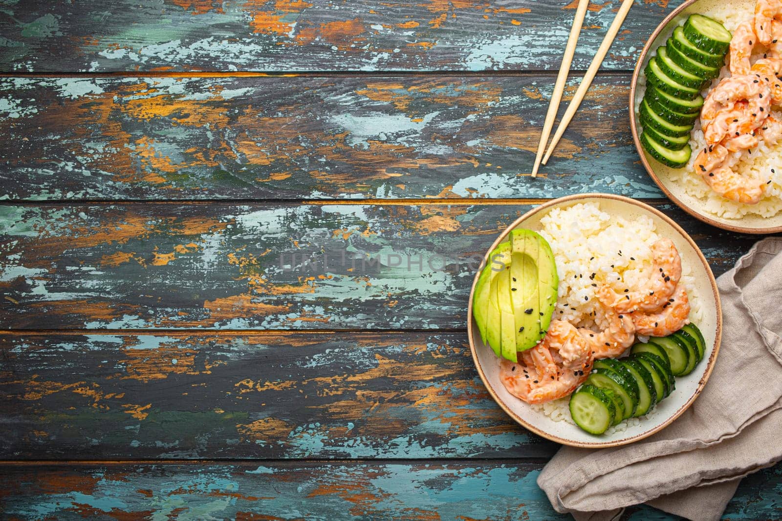 Two white ceramic bowls with rice, shrimps, avocado, vegetables and sesame seeds and chopsticks on colourful rustic wooden background top view. Healthy asian style poke bowl, space for text.