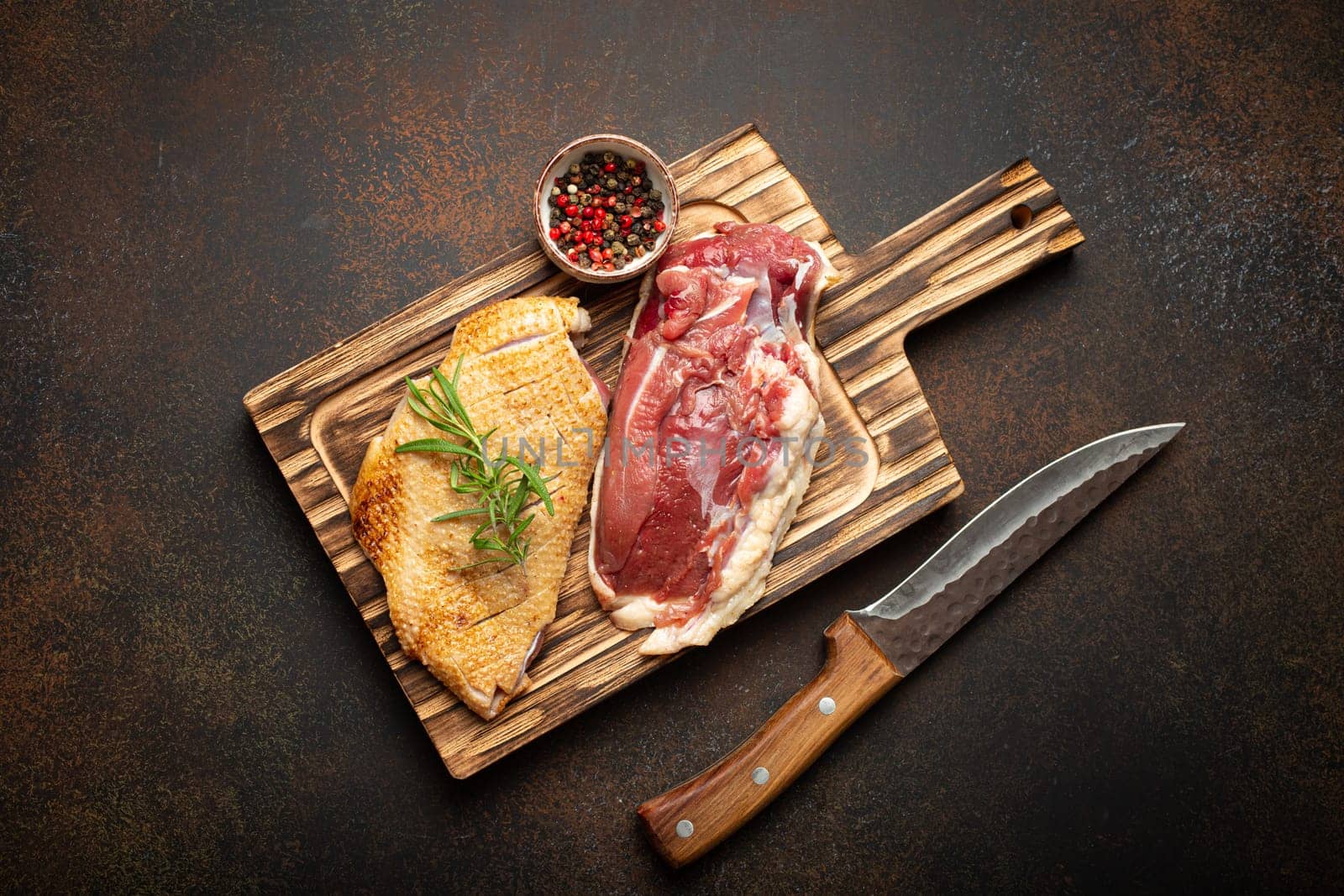 Two raw uncooked duck breast fillets with skin, seasoned with salt, pepper, rosemary top view on wooden cutting board with knife, dark brown concrete rustic background.