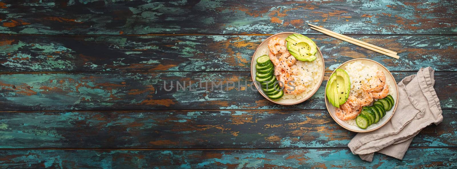 Two white ceramic bowls with rice, shrimps, avocado, vegetables and sesame seeds and chopsticks on colourful rustic wooden background top view. Healthy asian style poke bowl, space for text.