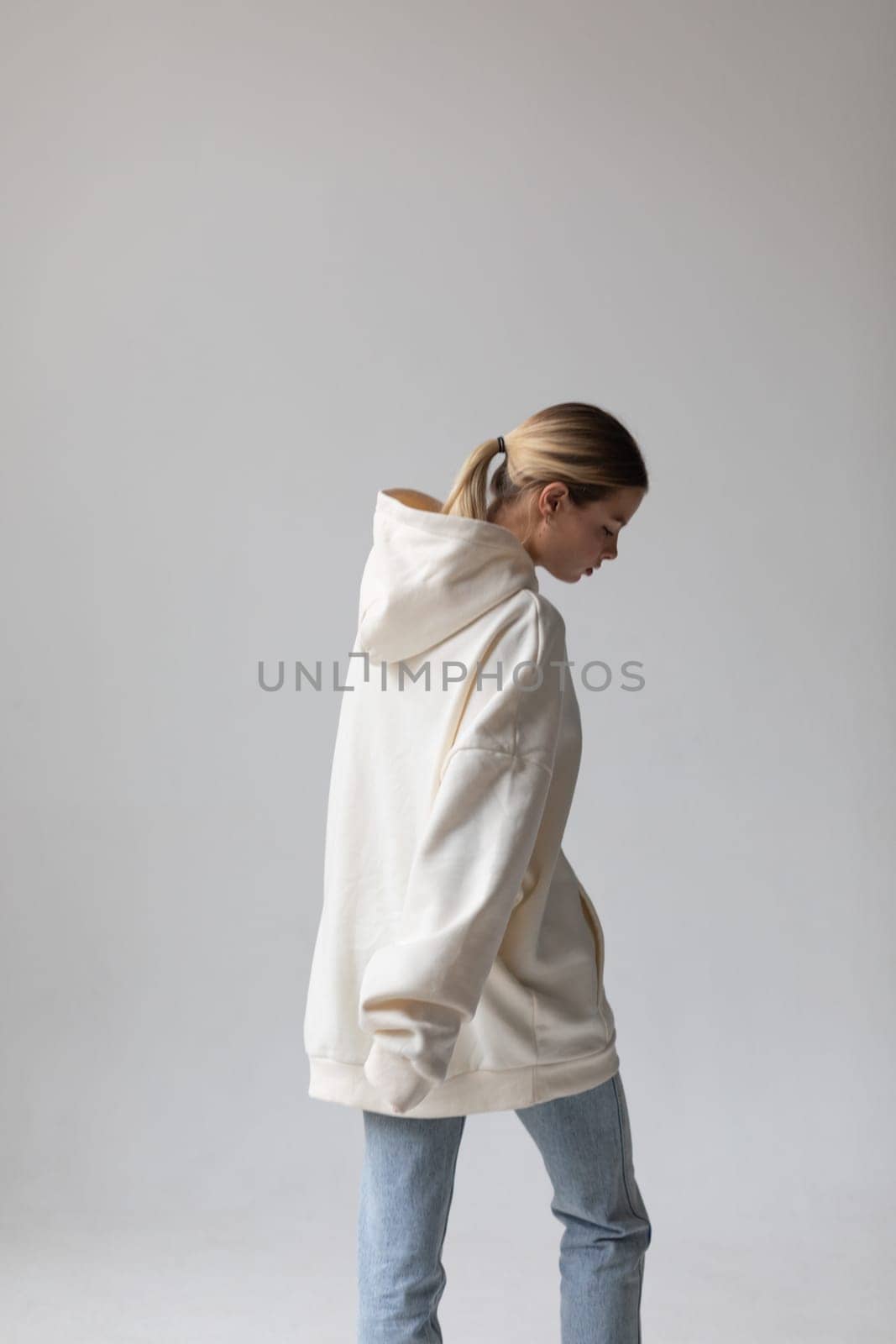 Beautiful blonde woman in a white hoodie and blue jeans posing on a white background by Freeman_Studio