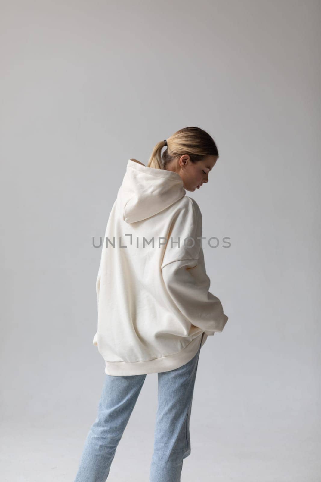 Beautiful blonde woman in a white hoodie and blue jeans posing on a white background by Freeman_Studio