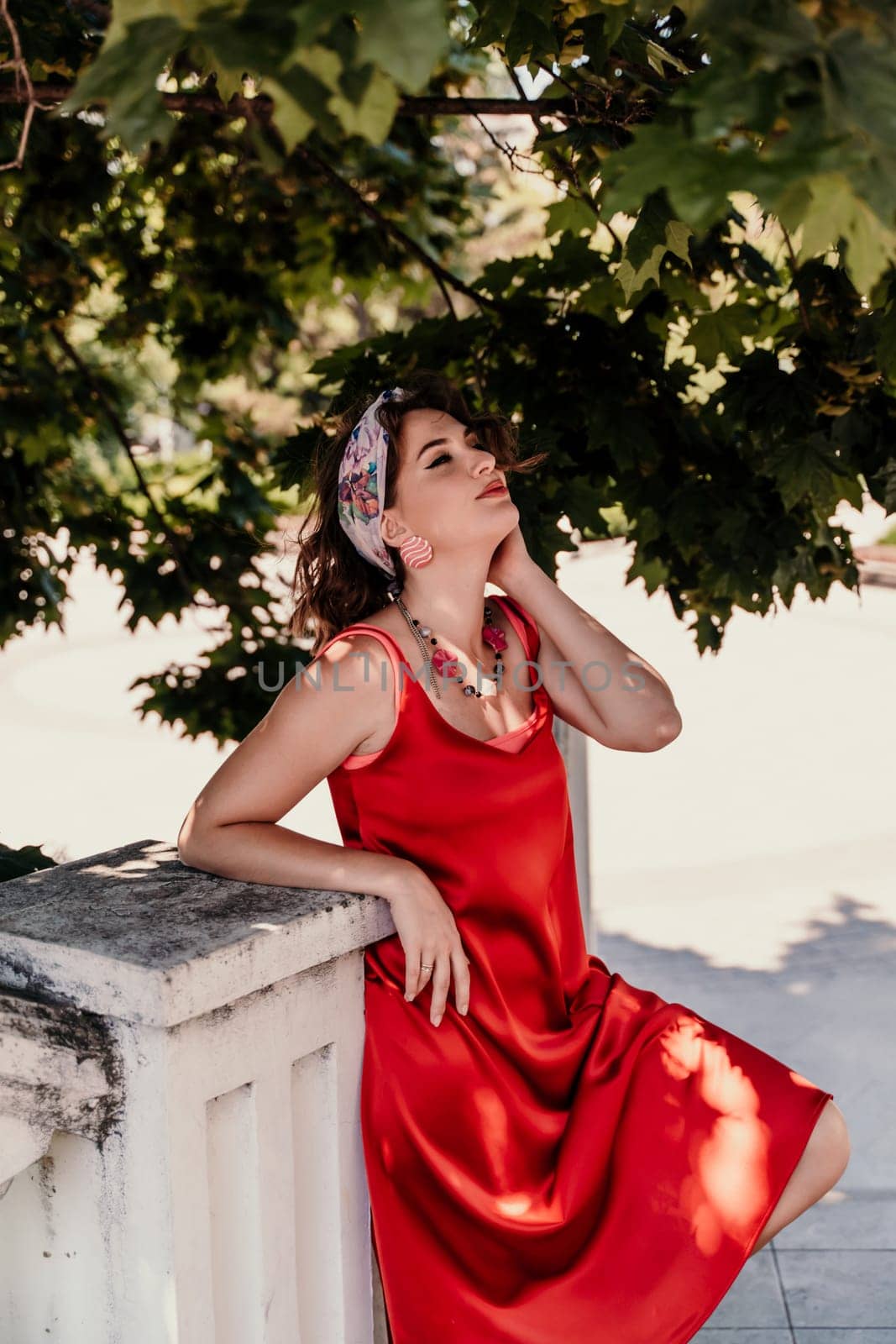 woman in a red silk dress and a bandage on her head smiles against the background of the leaves of a tree. She is leaning on the coop and looking into the camera. Vertical photo. by Matiunina