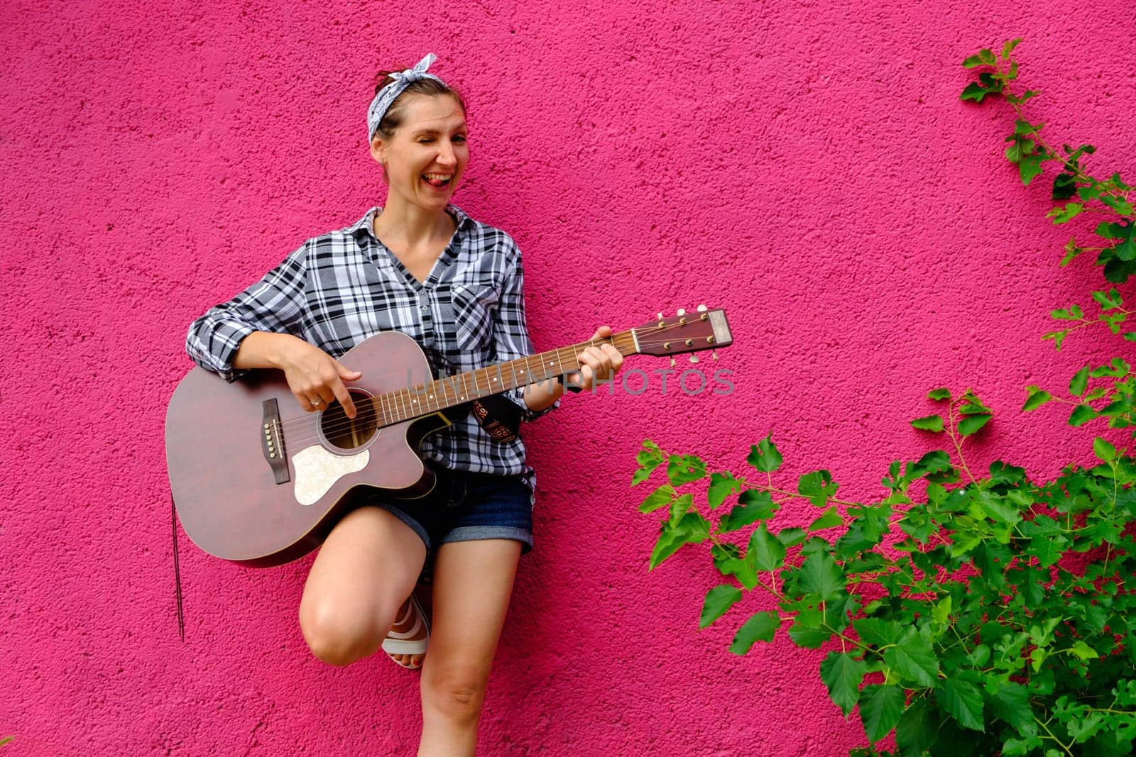 Young woman playing acoustic guitar on pink wall and green leaves background. Hipster girl portrait on street. Positive girl with guitar download