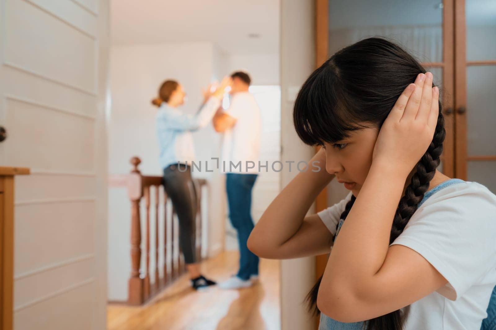 Stressed and unhappy young girl huddle in corner, cover her ears blocking sound of her parent arguing in background. Domestic violence at home and traumatic childhood develop to depression. Synchronos