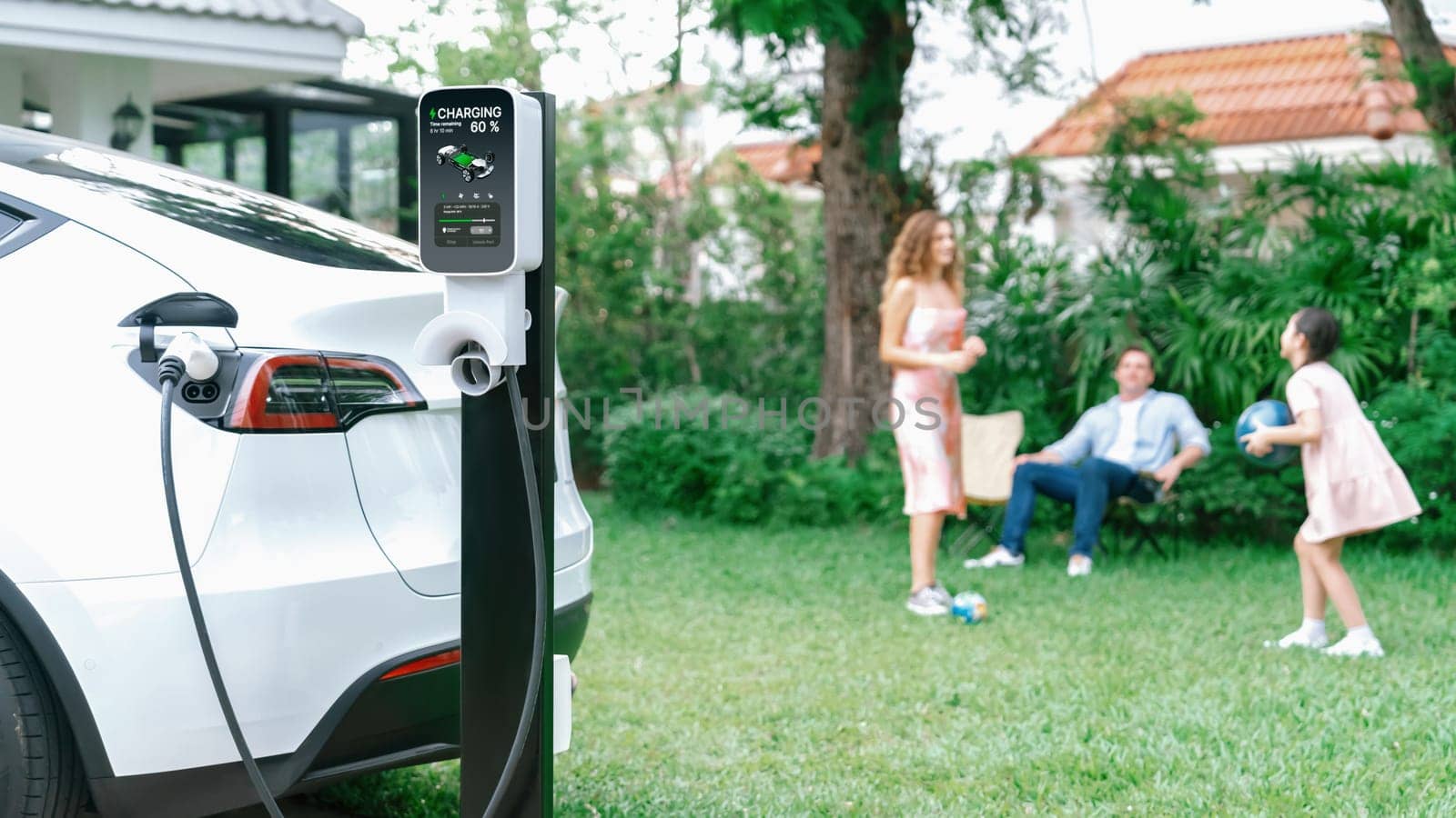 Focus electric vehicle recharge from home charging station on blur background of happy and playful family playing together. EV car using alternative and sustainable energy for better future.Synchronos