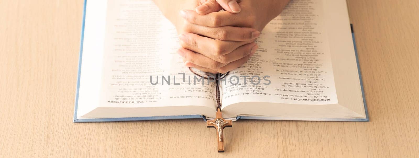 Cropped image of praying male hand holding cross on holy bible book at wooden table. Top view. Concept of hope, religion, faith, christianity and god blessing. Warm and brown background Burgeoning.