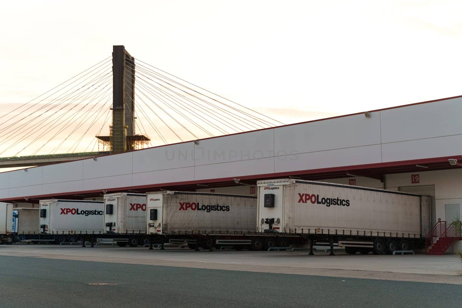 Seville, Spain - June 1, 2023: Several XPO Logistics trailers parked at a loading dock with a cable-stayed bridge in the background, captured at dusk.