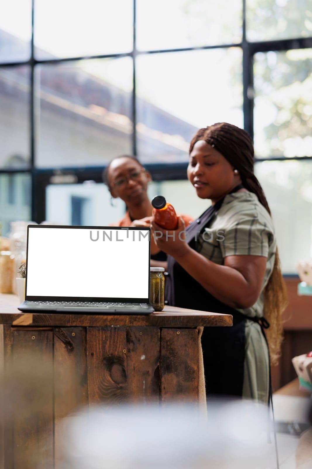 Wireless device with blank mockup template is placed on counter while shopkeeper showcases organic product in bottle. Digital laptop with an isolated white screen for advertisements.