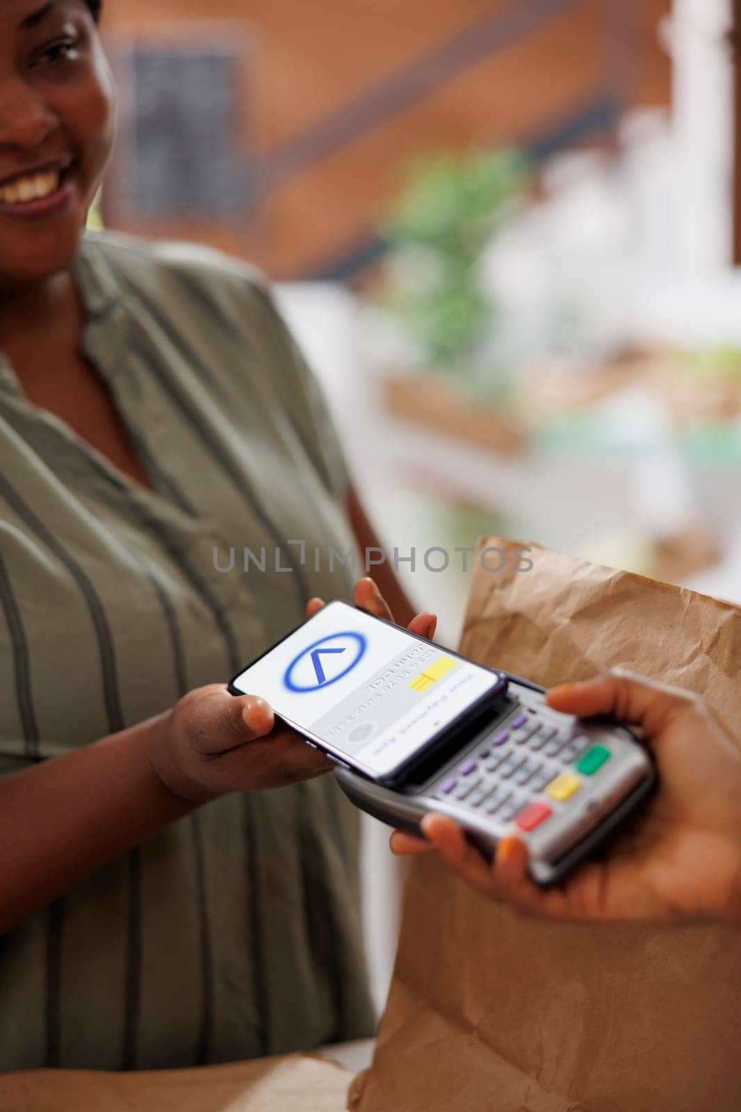 An African American woman smiles as she pays for freshly harvested organic produce using her mobile phone at a local marketplace. Convenient and eco-friendly shopping with contactless technology.