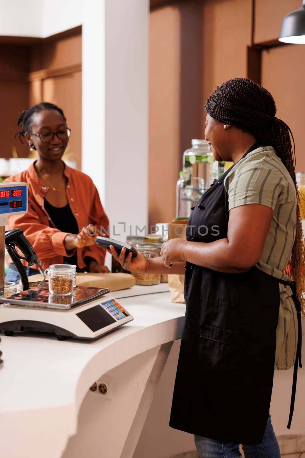 African American female client smiles while using her credit card for cashless payment. The local food market focuses on locally grown organic produce and eco friendly practices.