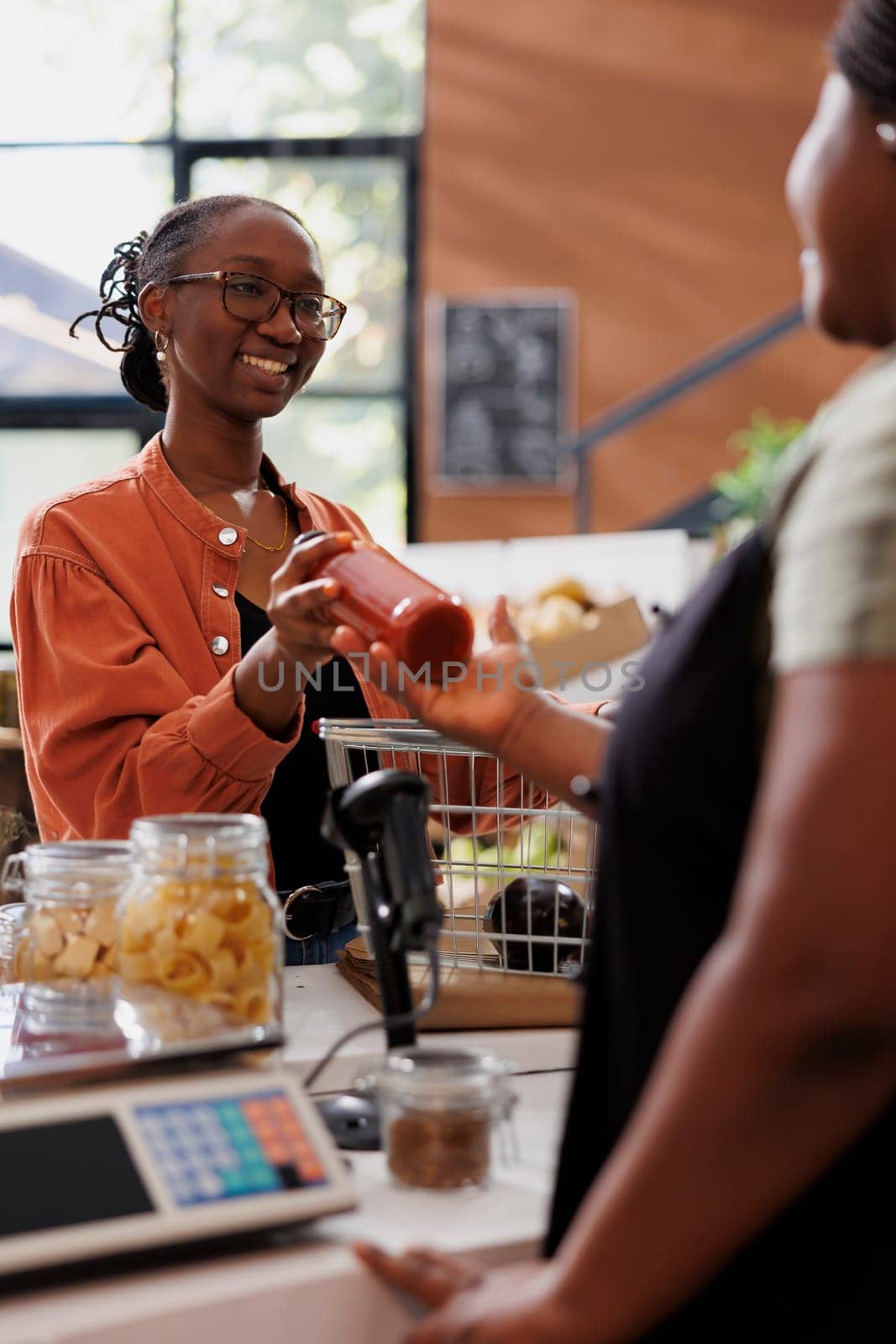 An African American woman sells freshly harvested organic produce to a customer in a grocery market. Female vendor receiving bottled sauce from smiling customer a checkout counter.