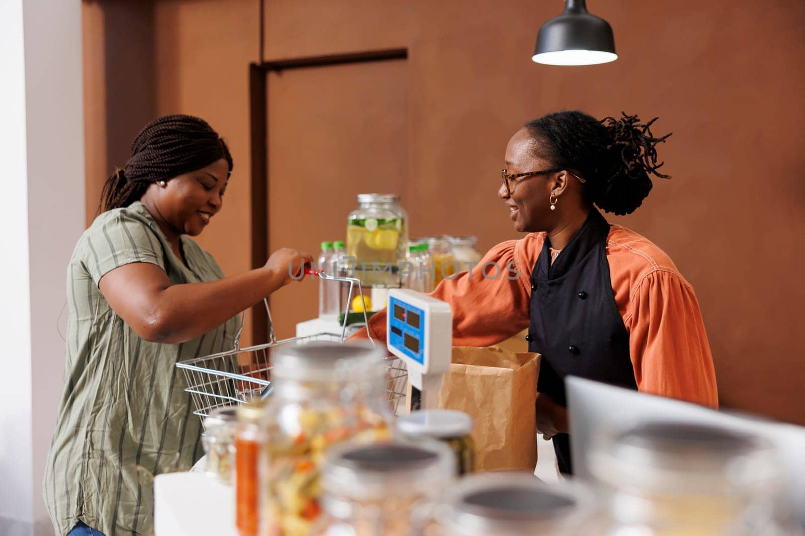 African American female customer unloading her shopping basket at the cashier counter. Friendly black woman vendor with a smile, assisting shopper with her farm grown food groceries.