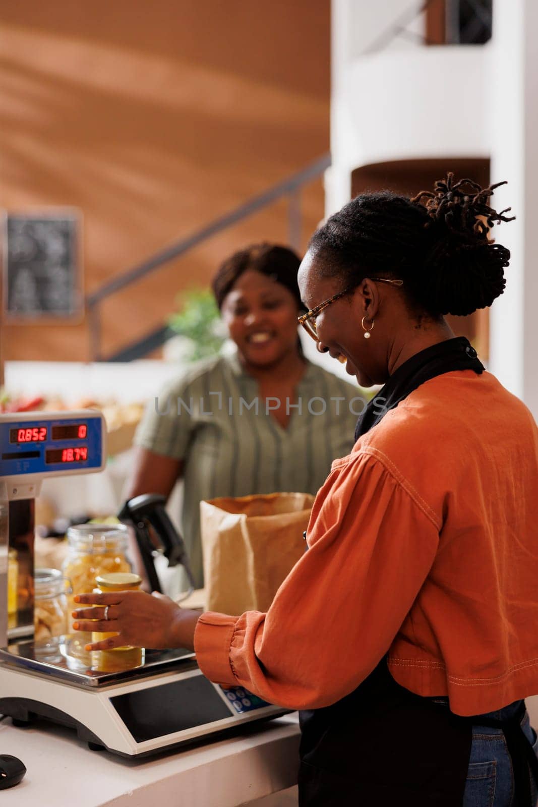 Female customer at a grocery store checkout counter with a smiling storekeeper weighing products on digital scale. Friendly vendor measuring weight of bio food items while client waits at desk.