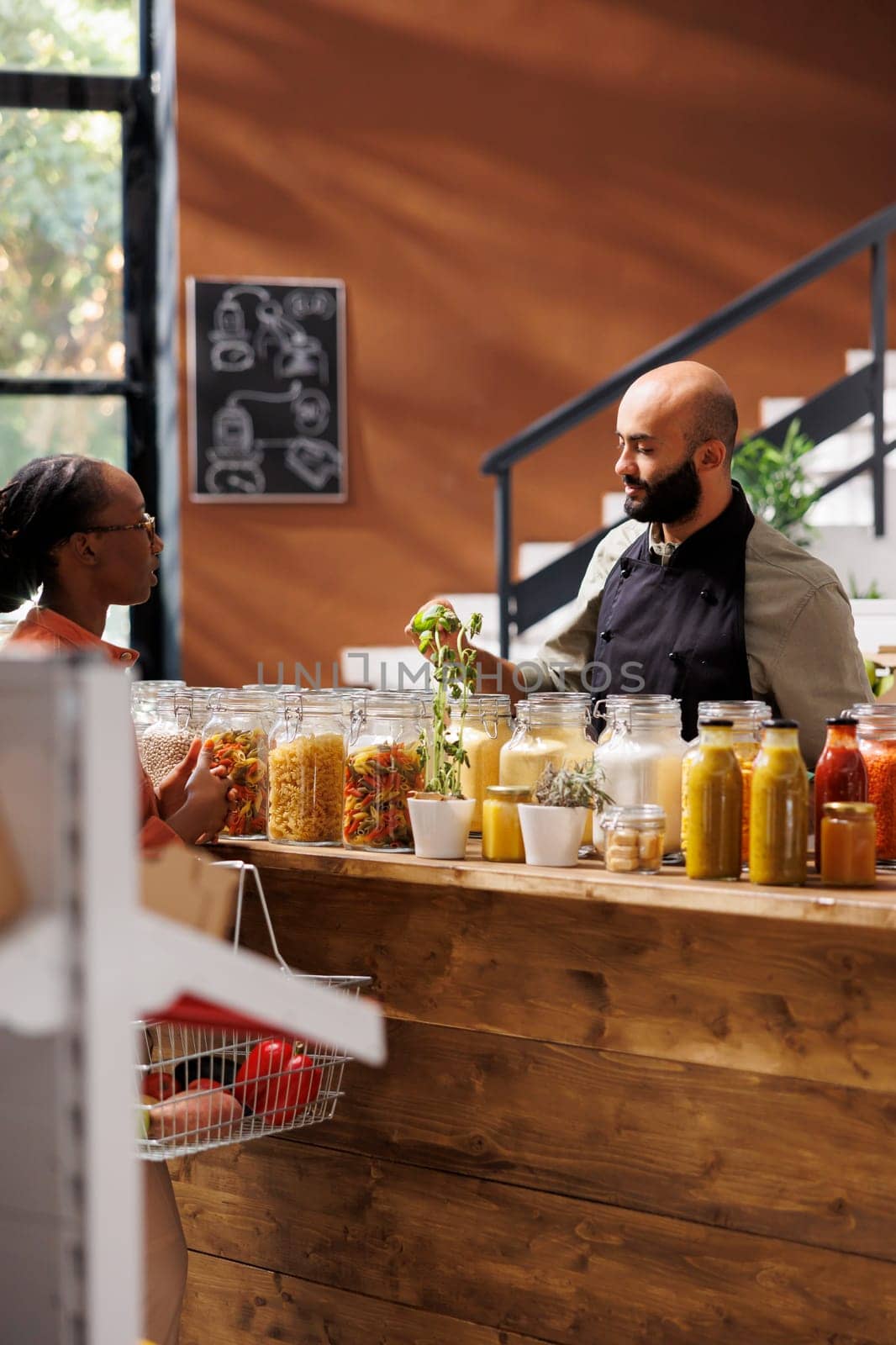 Black woman holding a basket filled with organic food products, in conversation with a male store worker. Male vendor advising african american customer with various sustainable produce.