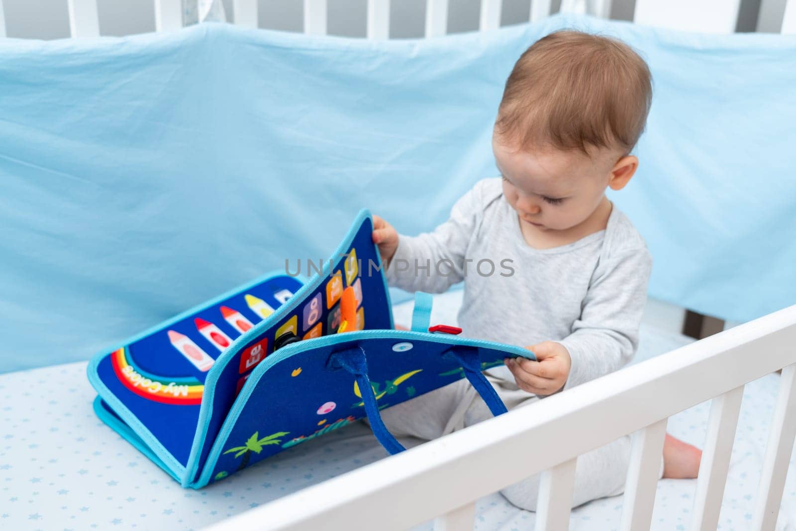 Baby playing with montessori busy book sitting in crib. Concept of smart books and modern toys by Mariakray