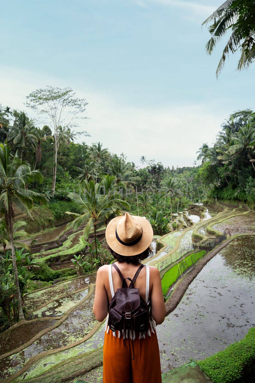 Back view of female tourist enjoying Tegalalang rice terrace landscape in Bali, Indonesia. Woman enjoying vacation and freedom in nature. Copy space. Vertical. Trip, vacation, mental health and healthy lifestyle concept.