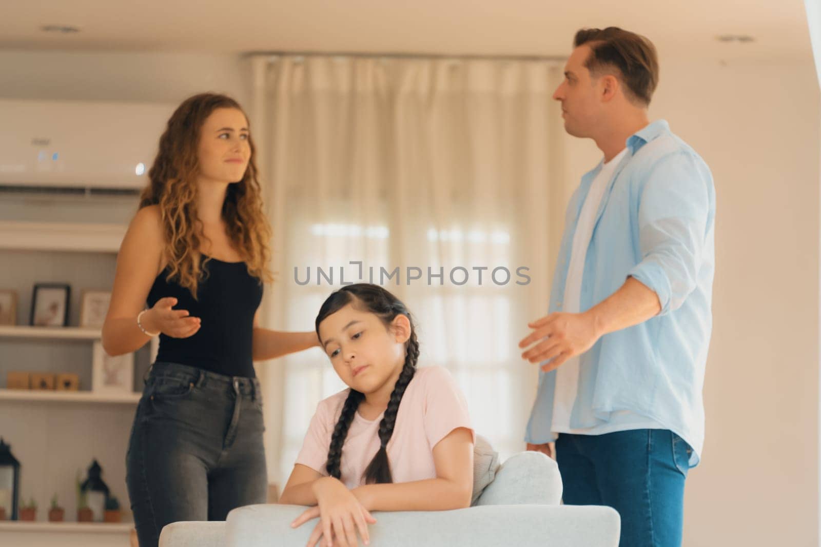 Annoyed and unhappy young girl sitting on sofa trapped in middle of tension by her parent argument in living room. Unhealthy domestic lifestyle and traumatic childhood develop to depression.Synchronos