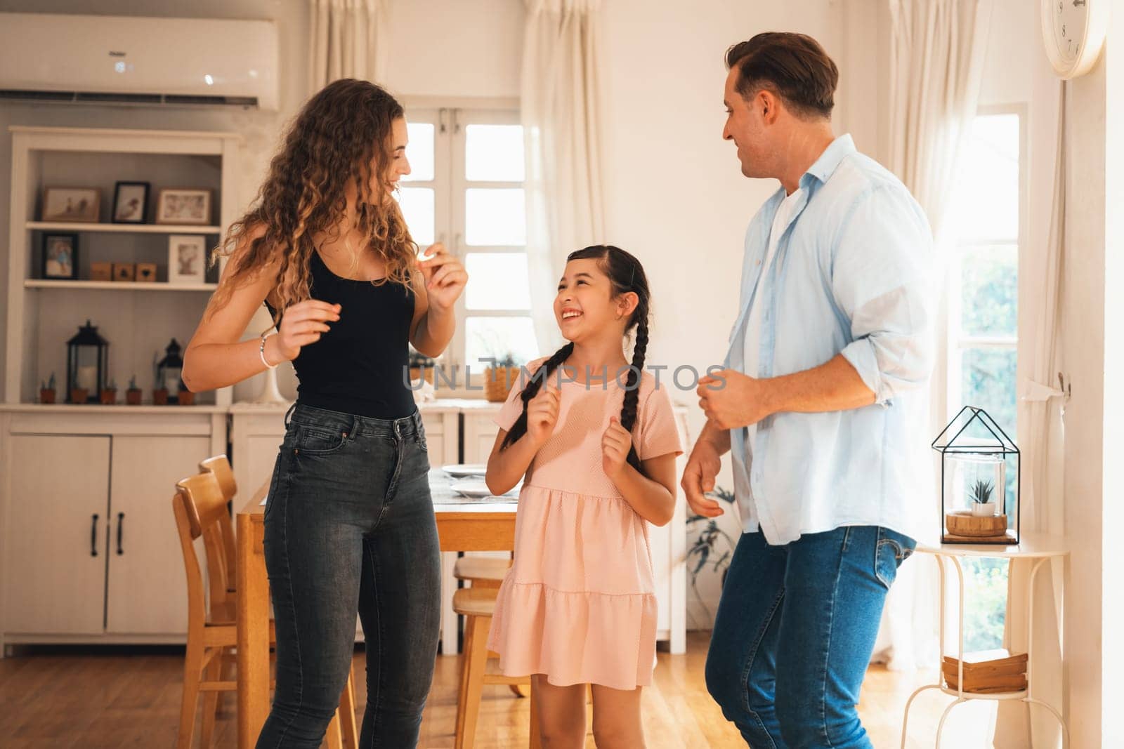 Happy family portrait with lovely little girl smiling and looking at camera, lovely and cheerful parent and their daughter sitting together in living room at home with warm daylight. Synchronos