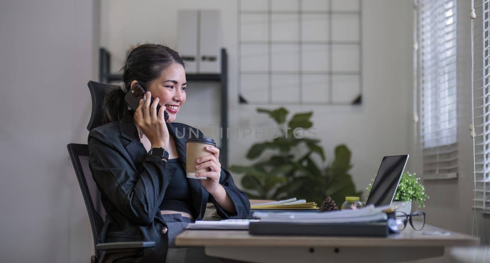 Young Asian business woman sits on the phone in an online business meeting using a laptop in a modern home office decorated with shady green plants..