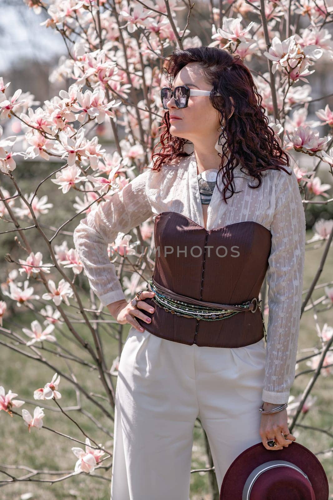 Magnolia park woman. Stylish woman in a hat stands near the magnolia bush in the park. Dressed in white corset pants and posing for the camera. by Matiunina