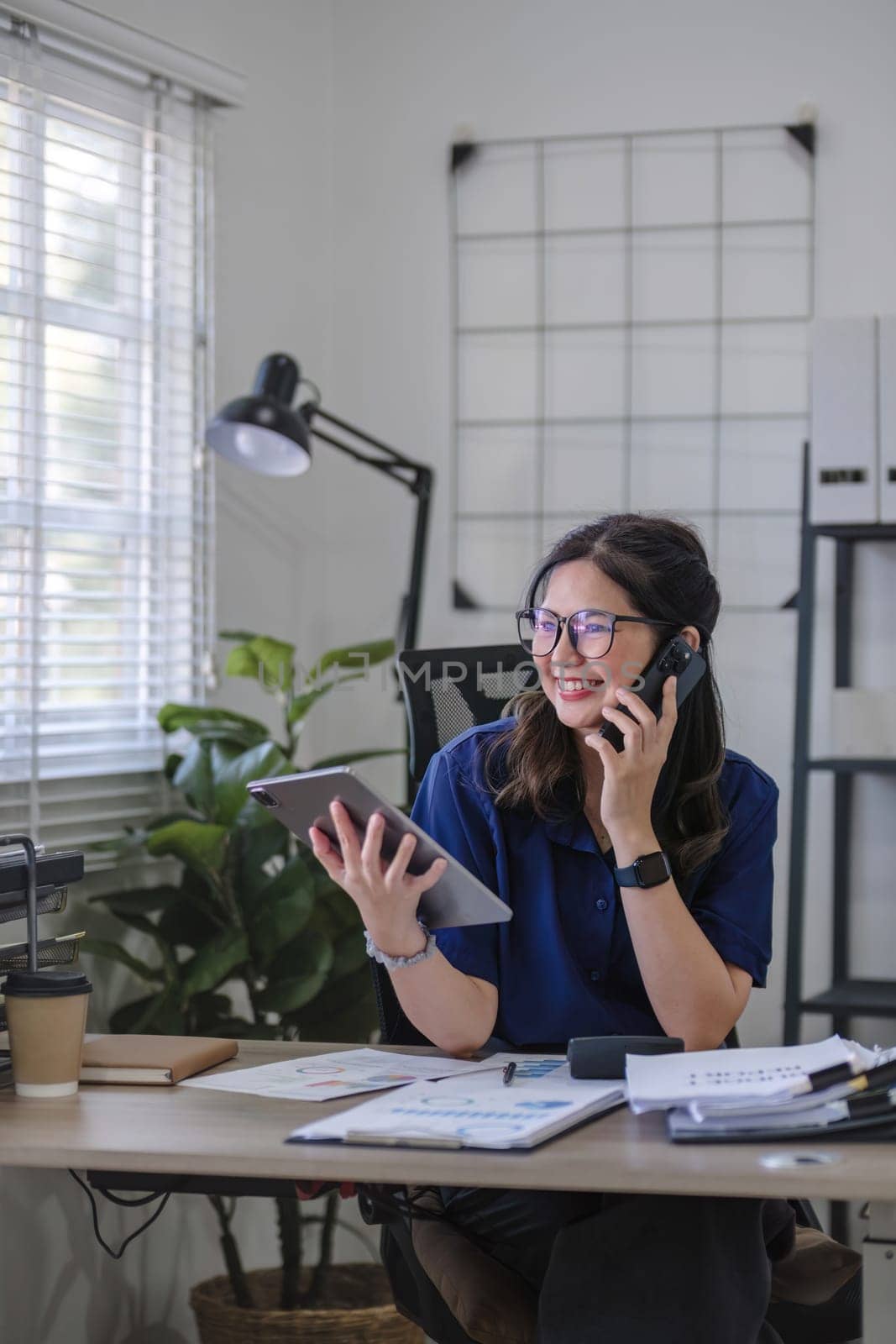 Young Asian business woman sits on the phone in an online business meeting using a laptop in a modern home office decorated with shady green plants. by wichayada