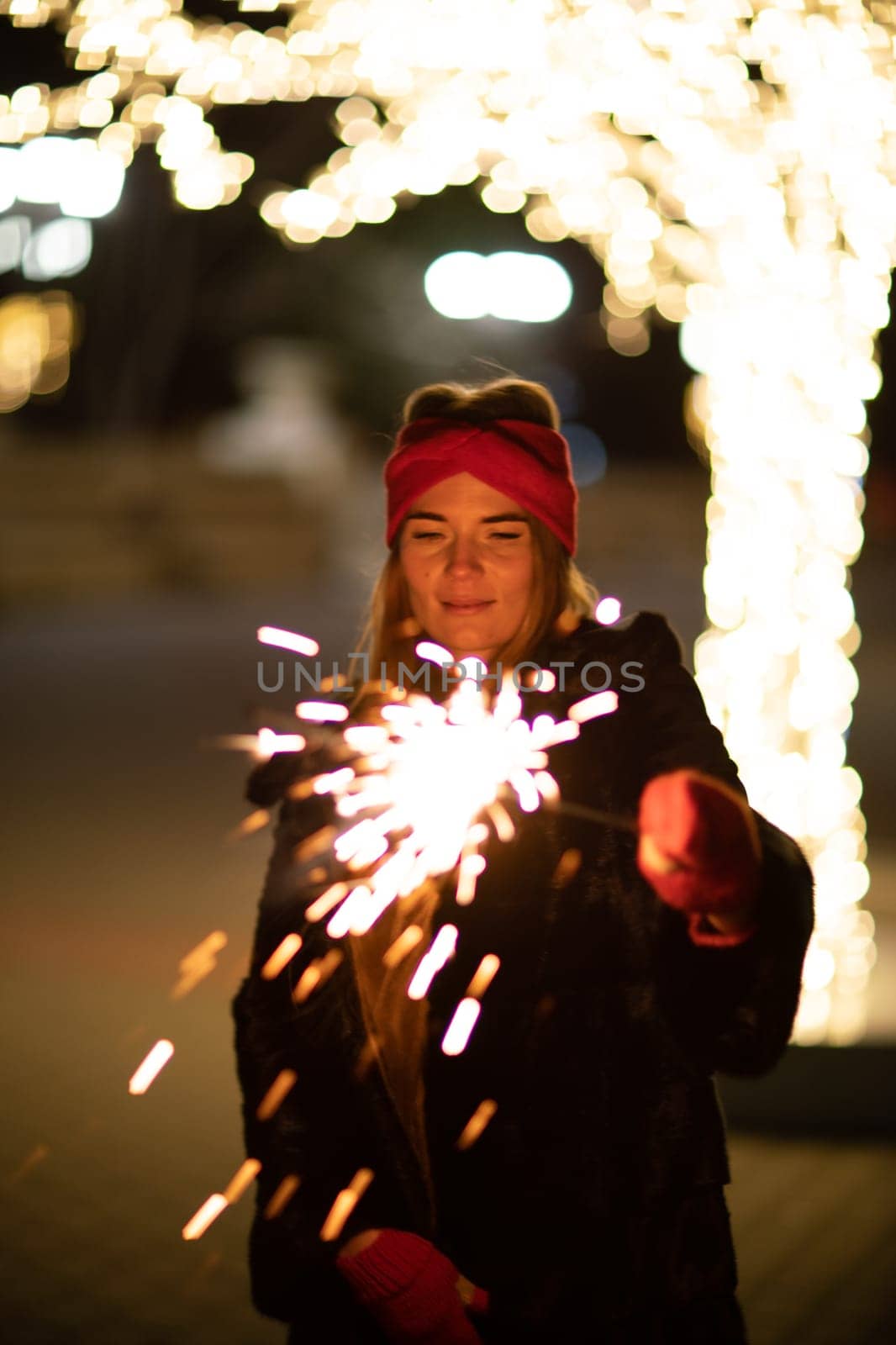 Woman holding sparkler night while celebrating Christmas outside. Dressed in a fur coat and a red headband. Blurred christmas decorations in the background. Selective focus by Matiunina