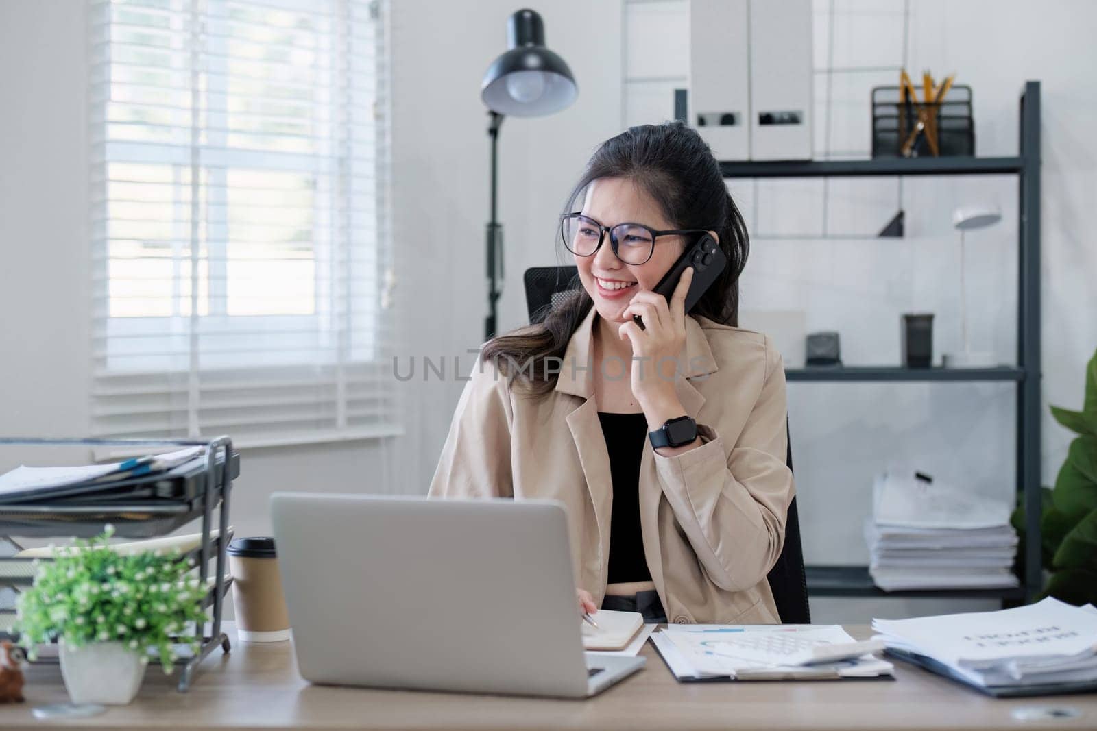 Young Asian business woman sits on the phone in an online business meeting using a laptop in a modern home office decorated with shady green plants..