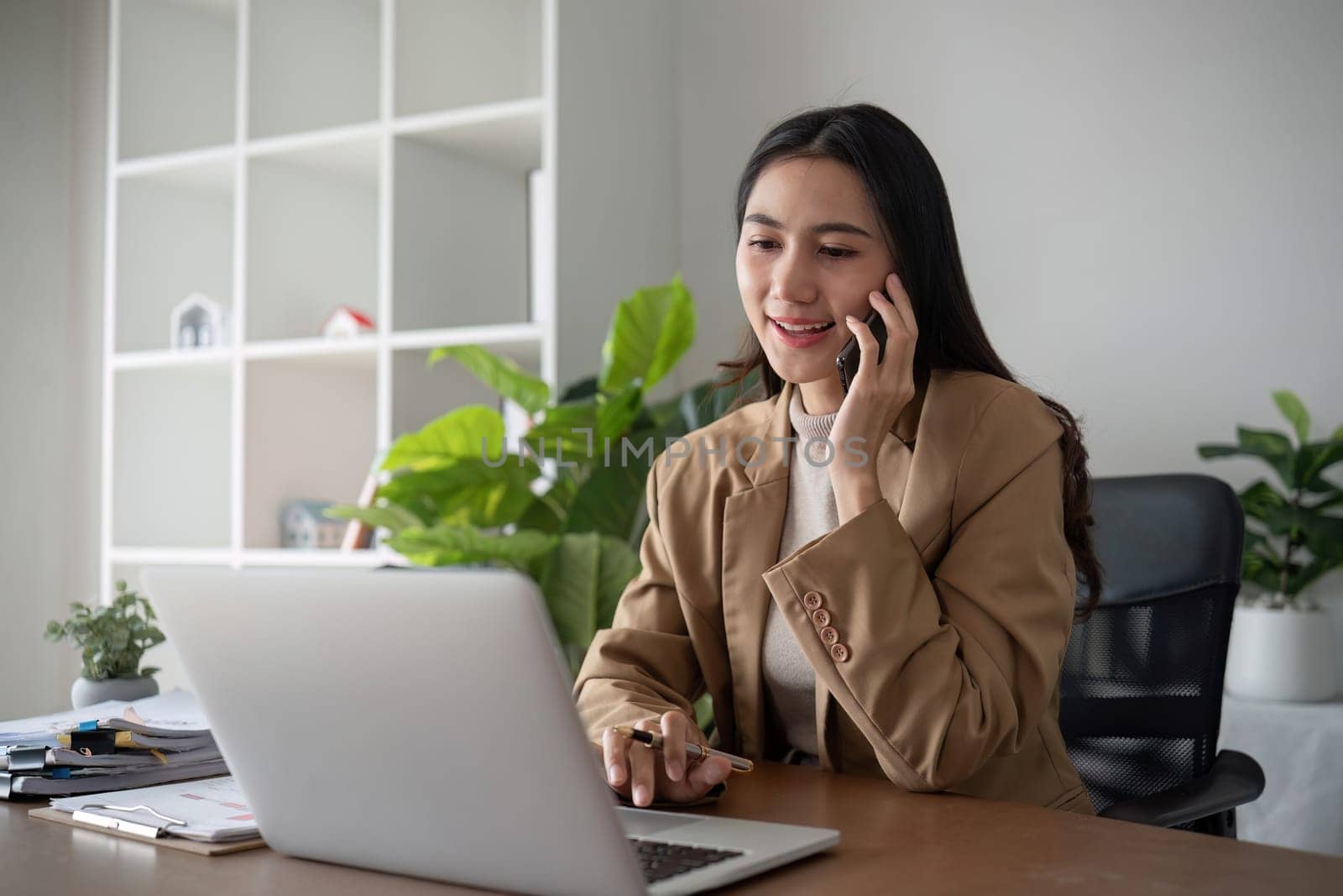 Young Asian business woman sits on the phone in an online business meeting using a laptop in a modern home office decorated with shady green plants..