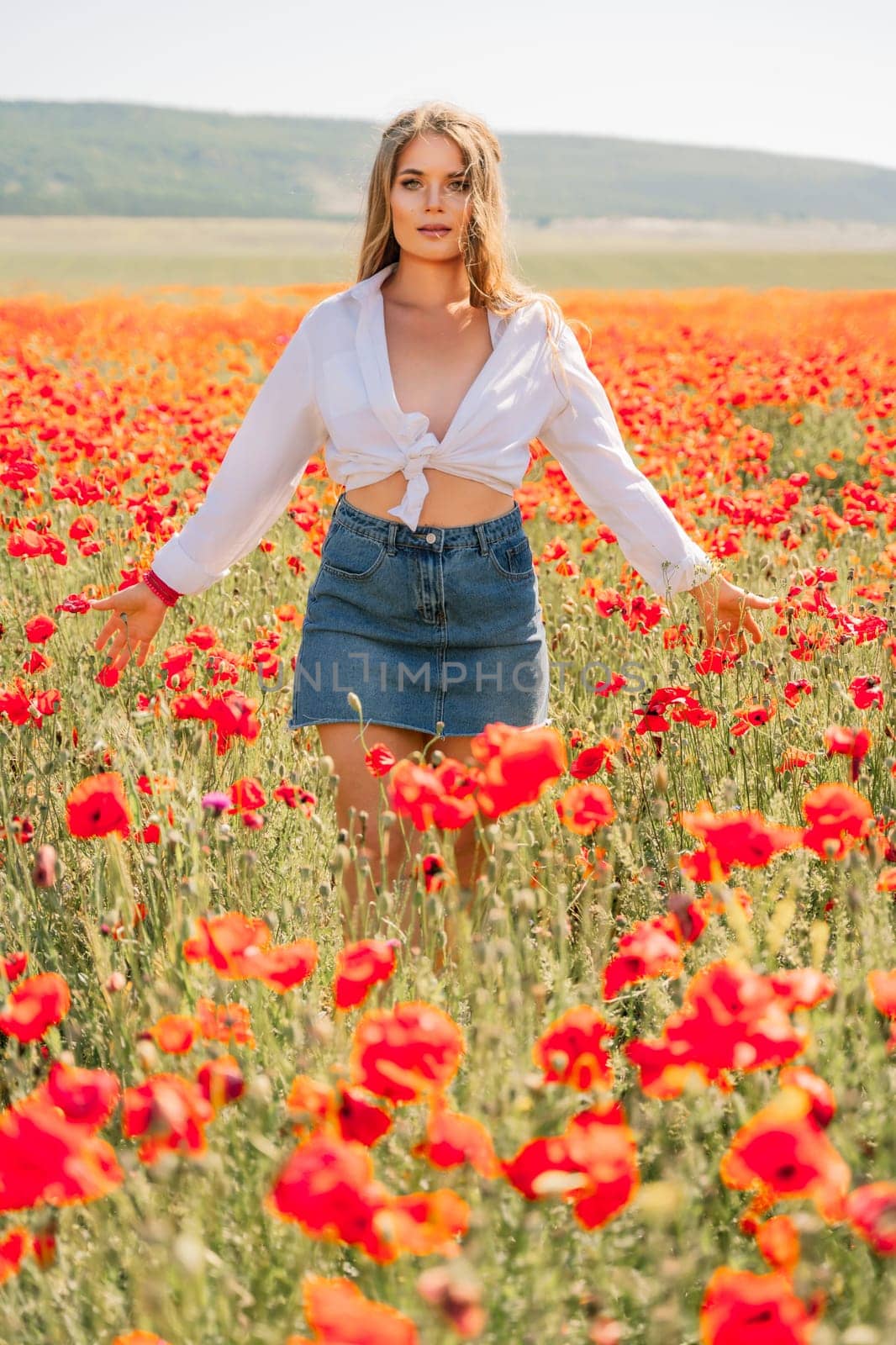 Happy woman in a poppy field in a white shirt and denim skirt with a wreath of poppies on her head posing and enjoying the poppy field. by Matiunina