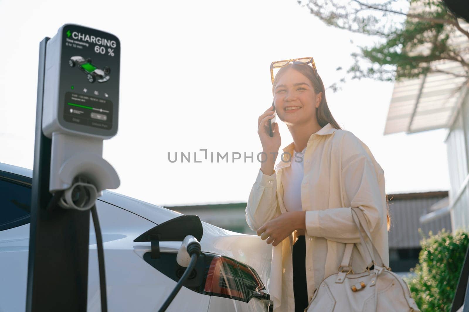 Young woman holding shopping bag talking on the phone while recharging EV car battery from charging station at city mall parking lot. Modern woman go shopping by eco car. Expedient