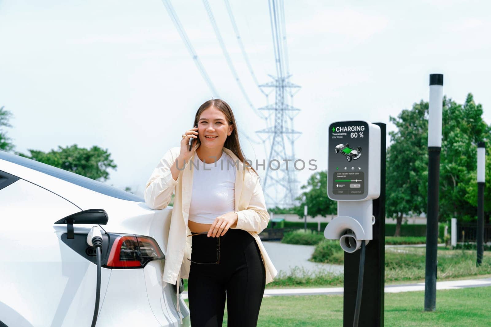 Young woman recharging EV car battery while talk on phone at charging station connected to electrical power grid tower facility as electrical industry for eco friendly vehicle utilization. Expedient