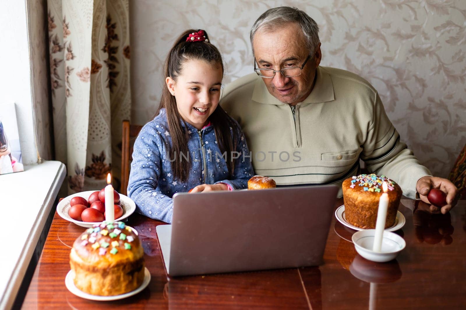 grandfather and granddaughter are talking via video link to their friends. Decorated table with colorful eggs and cake. Chatting during the COVID pandemic and the Easter holidays.