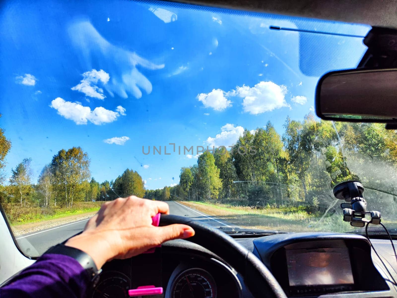 view from a car windshield of natural landscape with road, green trees and blue sky in summer or spring time. Hand of woman on steering wheel. Female traveler driving alone on trip or journey