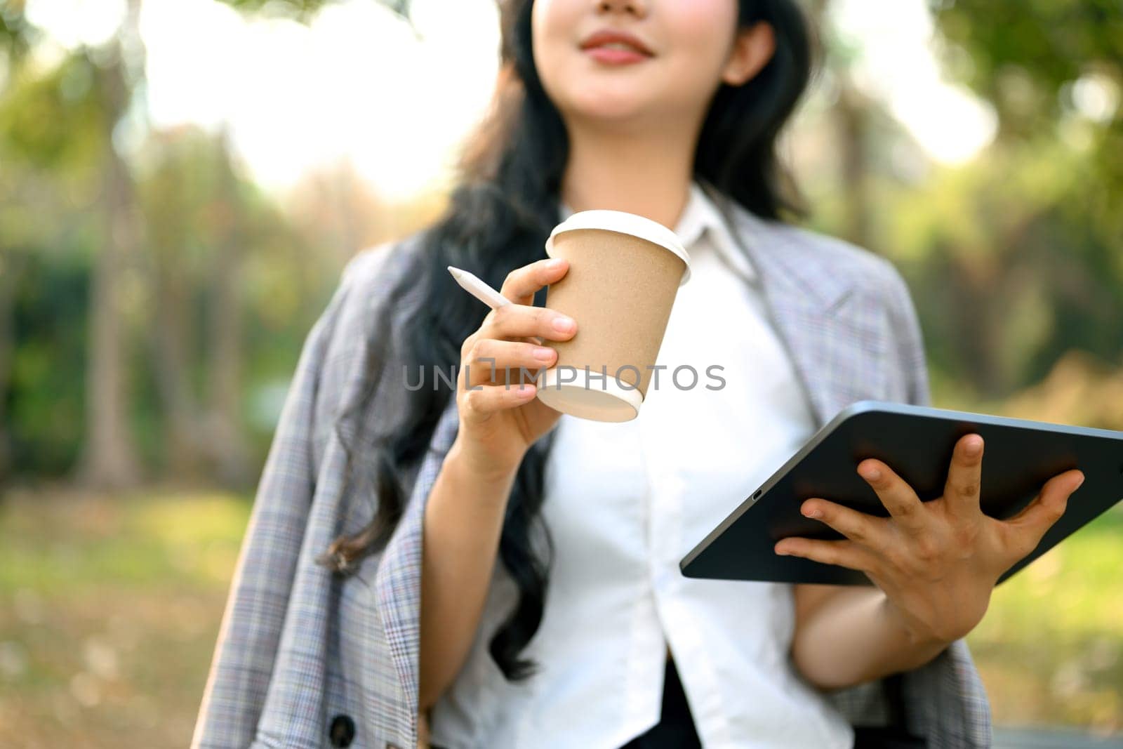 Satisfied young businesswoman drinking coffee and using digital tablet in the park.