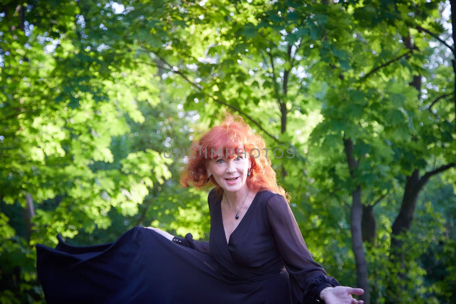 Elegant senior mature Woman dancing in Black Dress in green park. Woman with red hair posing on nature