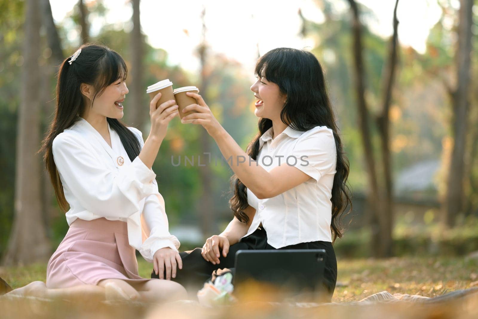Two cheerful female friends clinking paper cups with coffee and resting in the park after work.